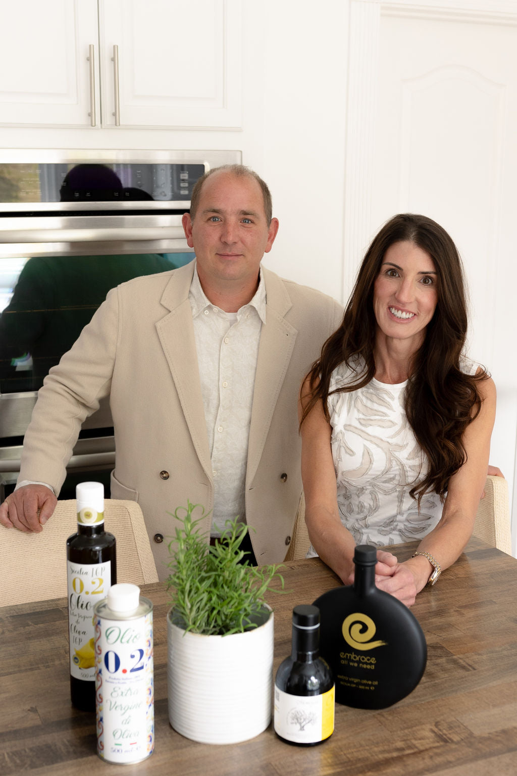 Two people standing behind a table with various bottles and a plant in a kitchen setting.
